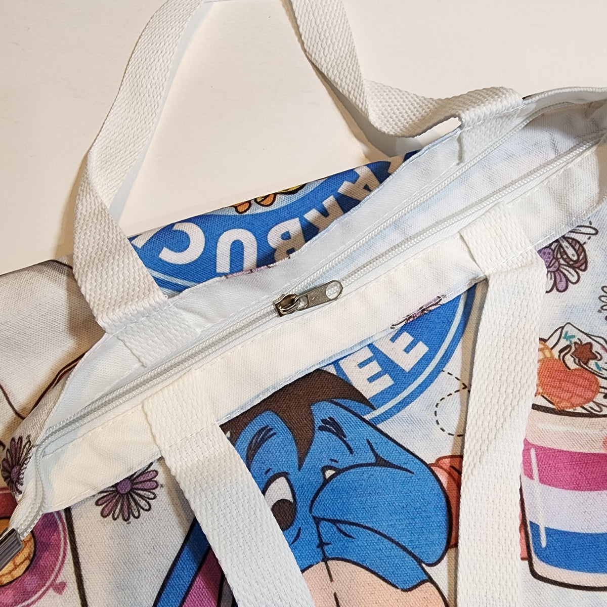 A close-up of a canvas tote bag with white webbing handles, a silver zipper, and a colorful cartoon print featuring a blue character and dessert illustrations.