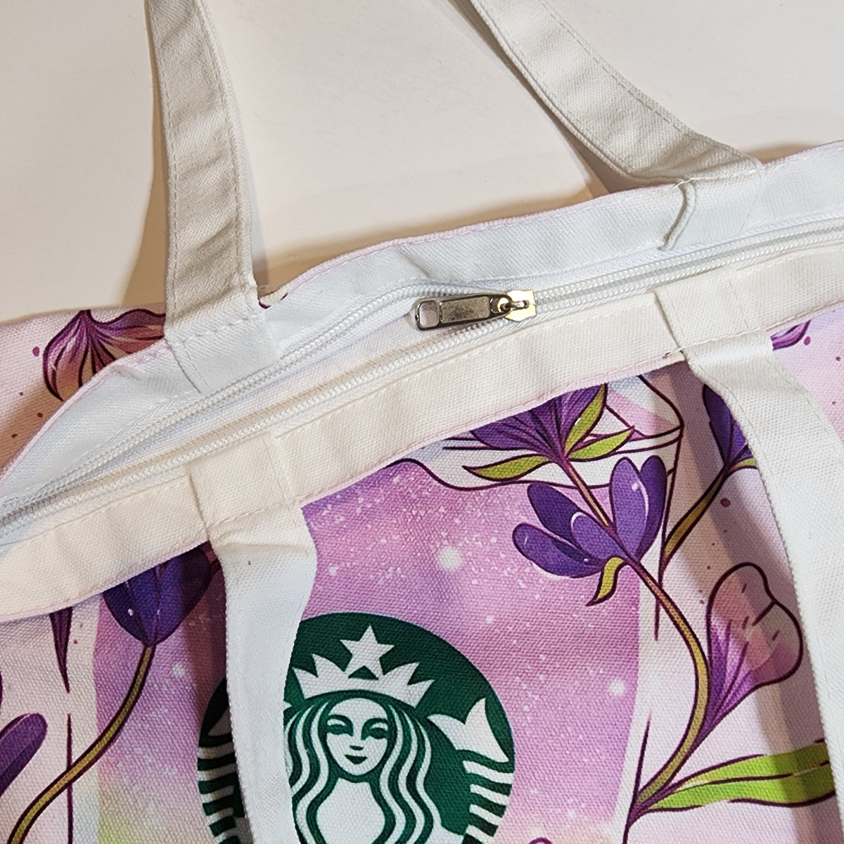 A close-up of a canvas tote bag with a pink and purple floral print, featuring a green Starbucks siren logo and a white zipper closure.