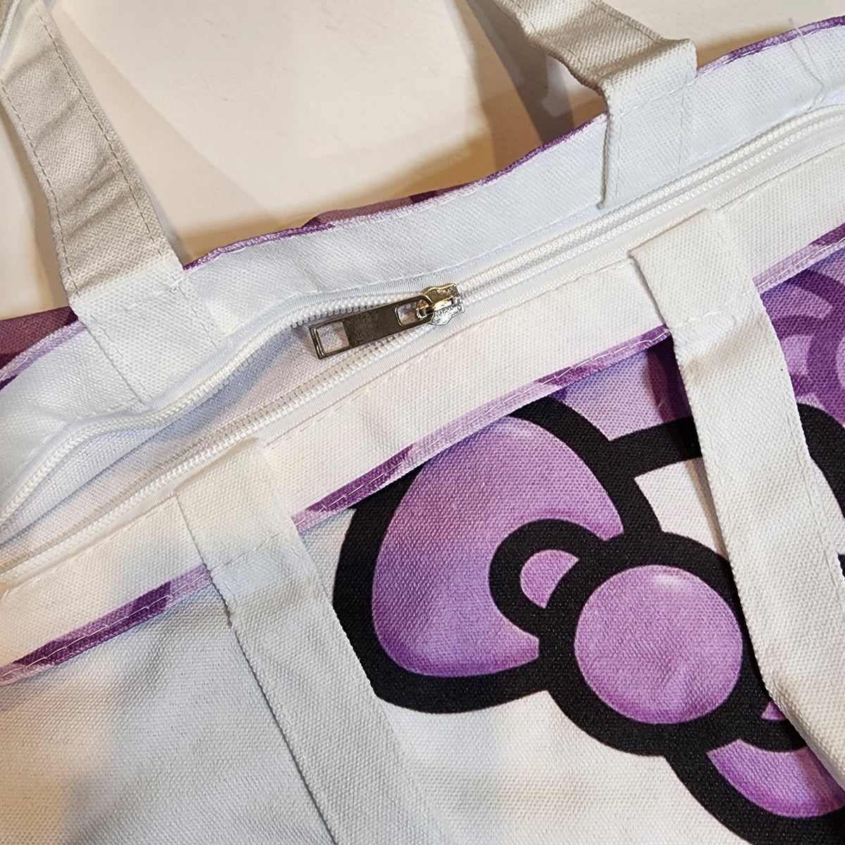 Close-up of a canvas tote bag with a white zipper, white fabric handles, and a large purple Hello Kitty bow graphic outlined in black.