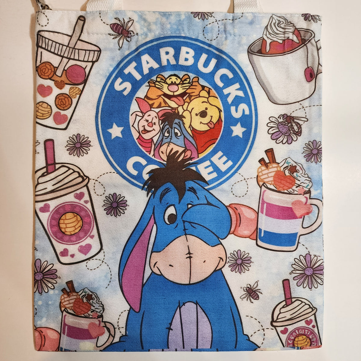 A fabric tote bag featuring a light blue watercolor background printed with a Disney Winnie the Pooh theme. The design includes a large blue Eeyore in the center, a large blue Starbucks Coffee logo with character illustrations, and multiple coffee cup illustrations with hearts and flowers.