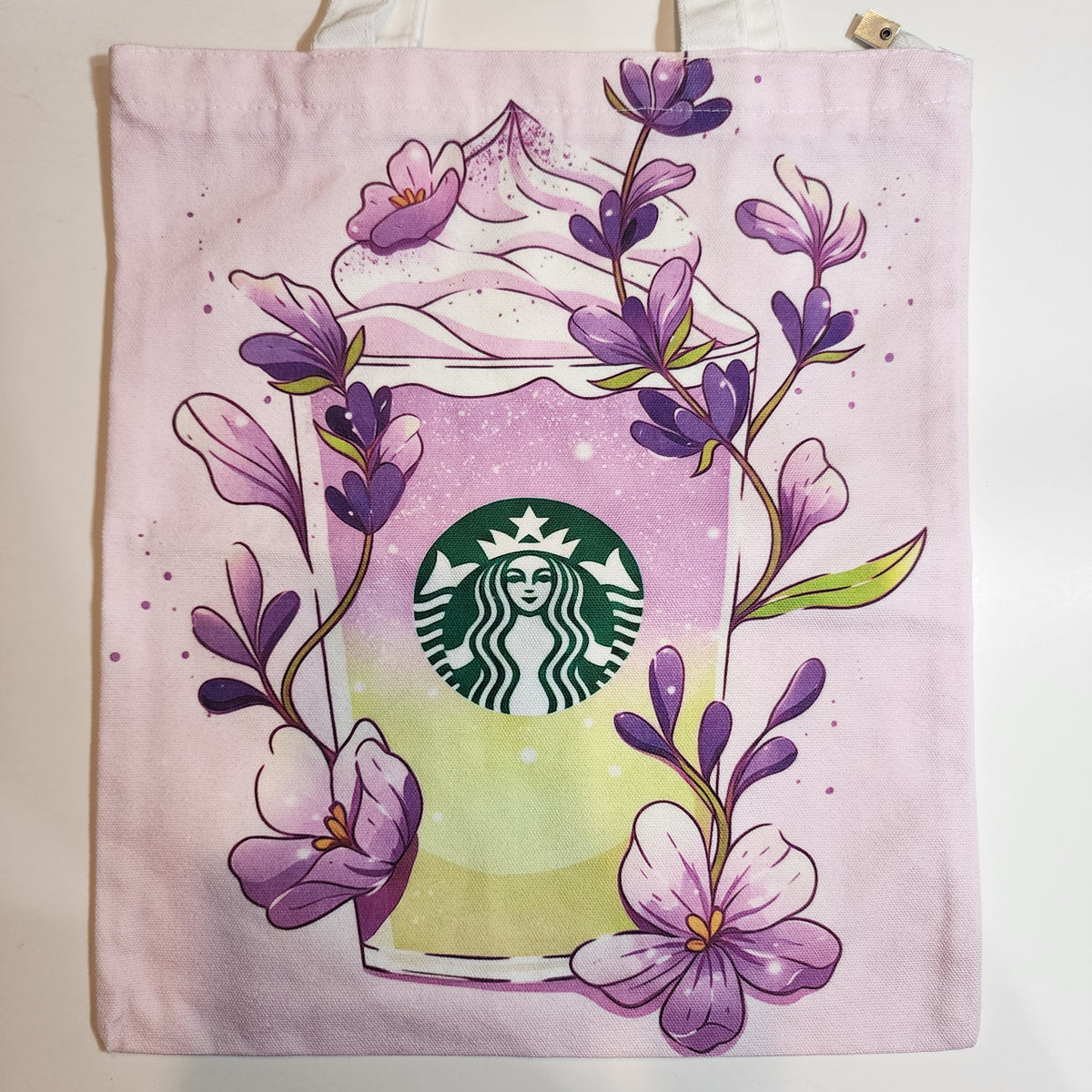 A light pink canvas tote bag featuring a printed illustration of a Starbucks coffee drink with purple flowers and green leaves.