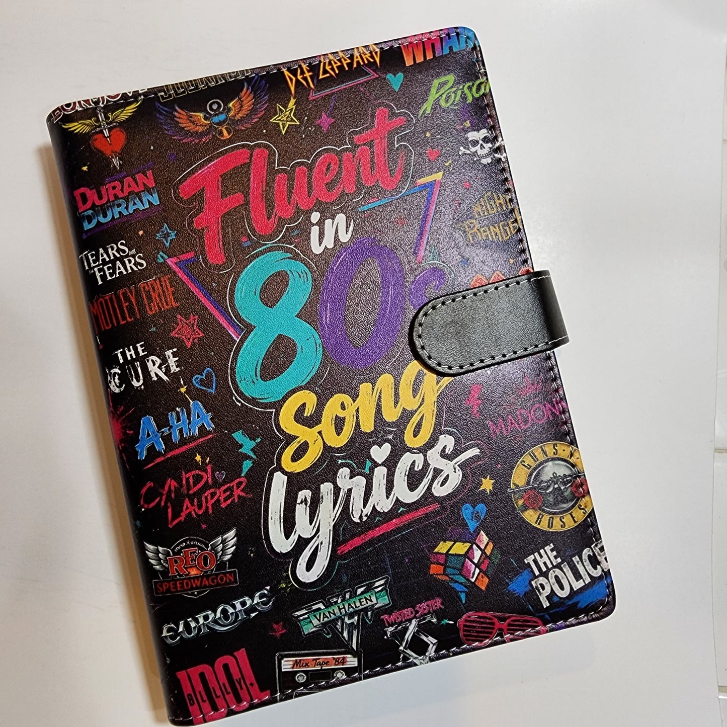 A black faux-leather passport cover featuring a colorful 1980s song lyrics collage design with various music artist names and graphics.