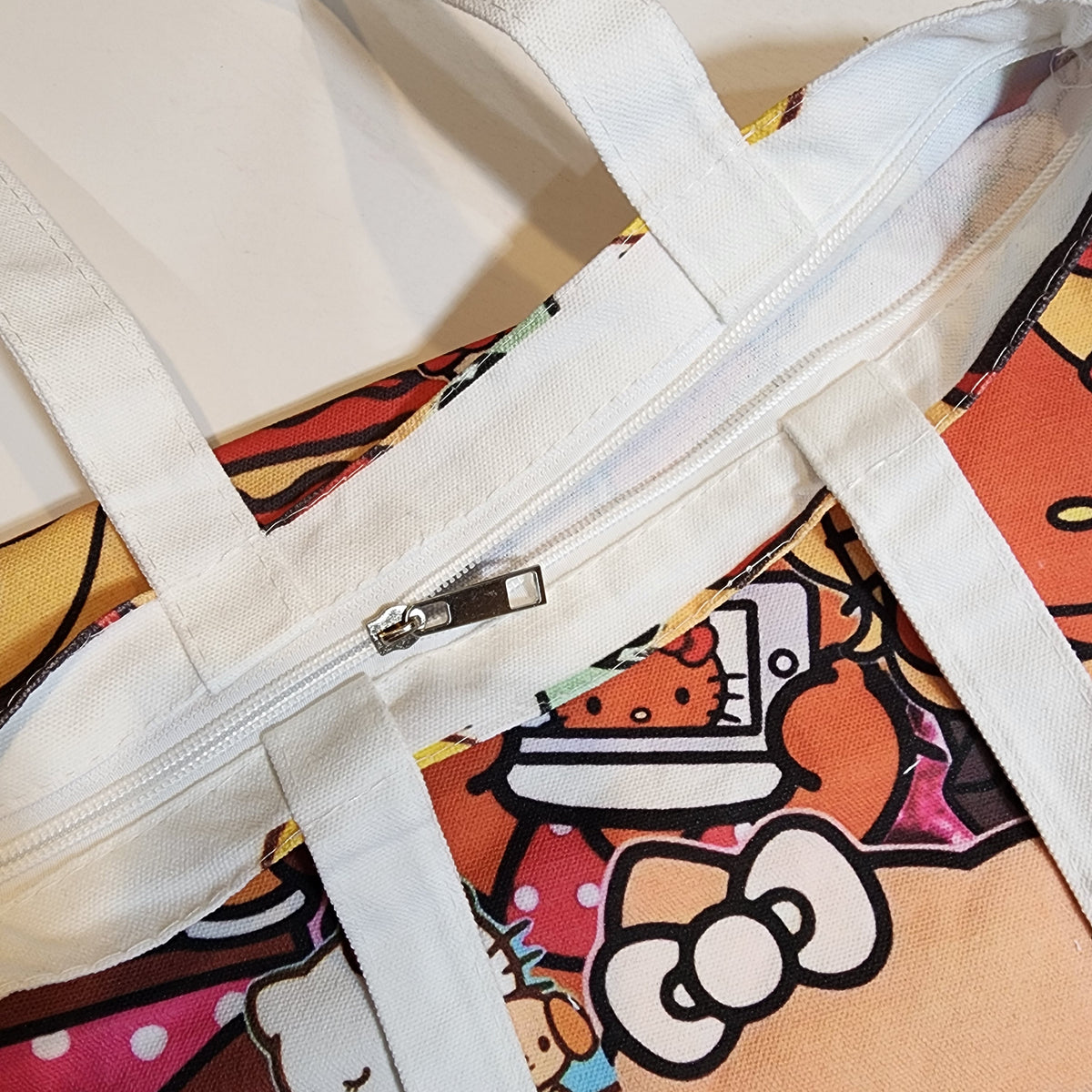 Close-up of a canvas tote bag with white handles and a colorful Hello Kitty print, showing a silver zipper closure.