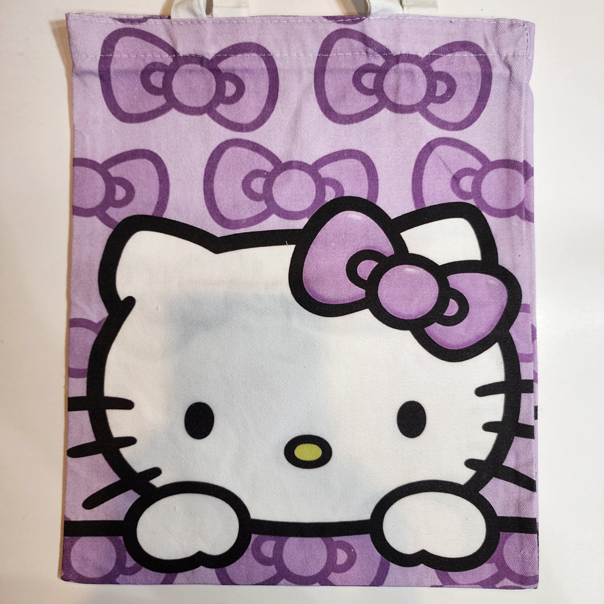 A light purple canvas tote bag featuring a large Hello Kitty face and bow on the front, with a repeating bow pattern in the background.